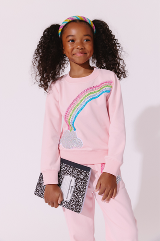 Lola and The Boys Rainbow Dreams Joggers Set
A young girl with curly black hair wearing a pink sweatshirt and matching sweatpants, holding a black and white speckled notebook. The sweatshirt features a colorful sequined rainbow and cloud design on the front.