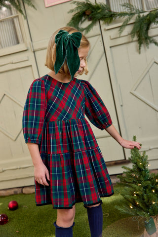 Pink Chicken Girls Maribelle Dress - Red Tartan
Young girl in a plaid dress with a green bow standing next to a small Christmas tree.