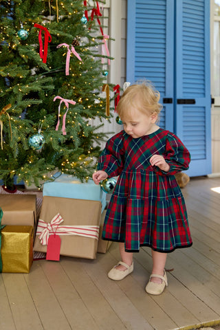 Pink Chicken Baby Girls Maribelle Dress Set - Red Tartan
Child in a plaid dress standing next to a decorated Christmas tree with presents underneath.