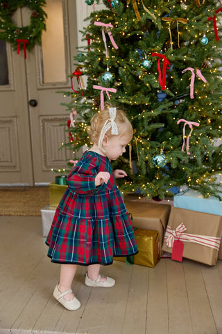Pink Chicken Baby Girls Maribelle Dress Set - Red Tartan
Child in a plaid dress standing in front of a decorated Christmas tree with presents underneath.