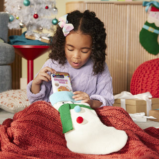 Magna-Tiles microMAGS Gingerbread House 24-Piece Travel Set
A young girl in a purple sweater sits under a red knit blanket, looking into a white Christmas stocking. The stocking has a green scarf and red and blue pom-poms, and she holds a package of Christmas cookies.