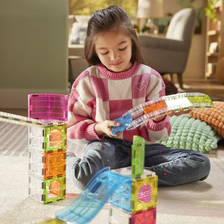 Magna-Tiles Rail Racers™ Max 80-Piece Set 
A young girl sits on a carpeted floor in a living room, playing with translucent magnetic building blocks. She is wearing a pink and white checkered sweater and blue jeans, holding a blue curved magnetic piece.