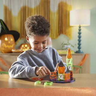 Magna-Tiles microMAGS Spooky 24-Piece Travel Set
A young boy wearing a gray sweatshirt sits at a wooden table, playing with a colorful magnetic building toy castle made of translucent orange, green, and black pieces. The background shows a cozy, softly lit room with Halloween decorations, including a jack-o'-lantern and a witch hat on a shelf, and a blue-green spiral lamp on a table lamp stand.