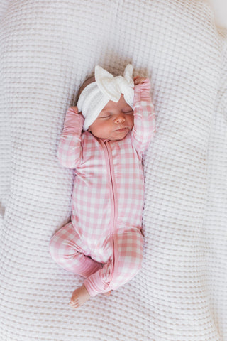 In My Jammers Blush Gingham Zipper Romper
Newborn baby in a pink checkered outfit with a white bow on a textured blanket