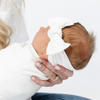 Lou Lou and Company Knit Headband - Finley
Person holding a baby wrapped in white fabric with a white headband.