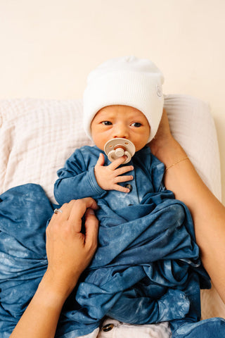 Lou Lou and Company Swaddle Blanket - Heath
Newborn baby wrapped in a blue blanket with a white hat and pacifier, held by an adult.