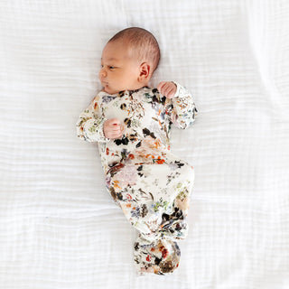 Lou Lou and Company Knotted Gown - Nellie
A newborn baby lying on a white textured blanket, wearing a long-sleeve footed sleeper with a multicolor floral and abstract print on a cream background.