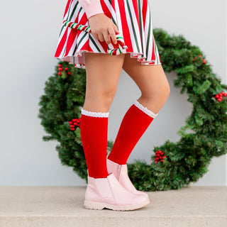 Little Stocking Co Lace Top Knee High Socks - Santa Baby
Person wearing a red and green striped dress with knee-high red socks and pink shoes in front of a Christmas wreath.