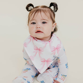 Copper Pearl Bandana Bib Set - Bow My Gosh
A baby wearing a white long-sleeve footed sleeper with pink bow and blue floral prints, along with a white bandana bib featuring pink bows, set against a plain light beige background.