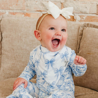 Copper Pearl Bandana Bib Set - Vintage Carnival
A baby wearing a blue and white toile-print one-piece jumpsuit and a matching bow headband, sitting on a beige couch.