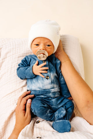 Lou Lou and Company Top and Bottoms - Heath
Baby in a blue outfit with a white hat and pacifier, held by an adult on a soft surface.