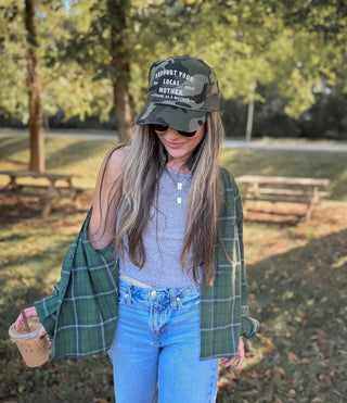 Support Your Local Mother® Trucker Hat - Camo
Woman wearing a cap and sunglasses, standing in a park with trees and benches in the background.