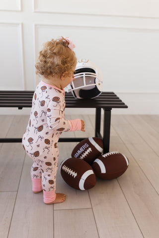 In My Jammers Blush Football Zipper Romper
Child in pajamas standing next to footballs and a helmet on a wooden floor.