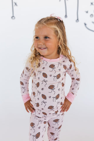In My Jammers Blush Football Two Piece Set
Young girl wearing a pink pajama set with football and bow pattern on a white background