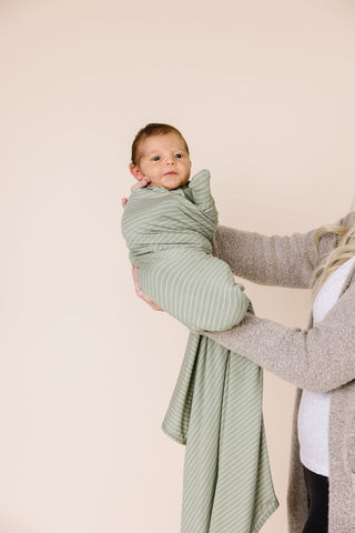 Lou Lou and Company Knit Swaddle Blanket - Carson
Baby wrapped in a green swaddle blanket held by an adult against a beige background