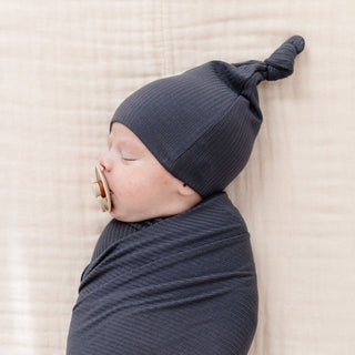 Lou Lou and Company Ribbed Top Knot Hat - Hudson
A sleeping newborn baby swaddled in a dark gray ribbed fabric wrap and wearing a matching pointed knit hat, lying on a textured beige blanket.