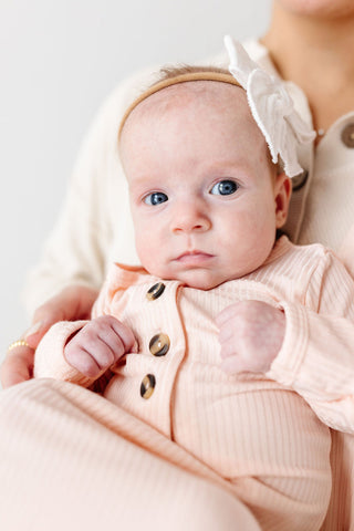 Lou Lou and Company Ribbed Knotted Gown - Rosie
Baby wearing a pink outfit with buttons and a headband, held by an adult.