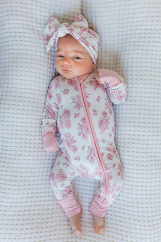 In My Jammers Blush Garden Zipper Romper
Newborn baby in a pink floral onesie and headband lying on a textured surface.