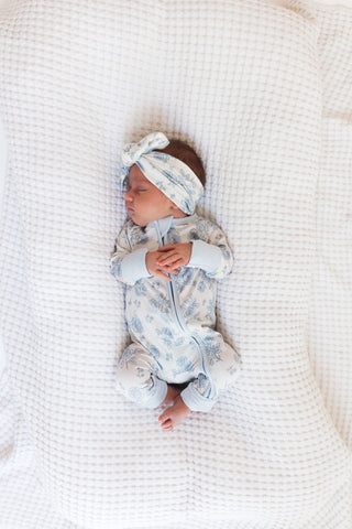 In My Jammers Blue Rose Garden Zipper Romper
Newborn baby in a floral outfit and headband lying on a white textured blanket.