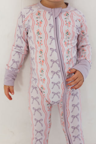 In My Jammers Zipper Romper - Purple Amelia Bow
Child wearing a purple onesie with floral and bow pattern on a white background