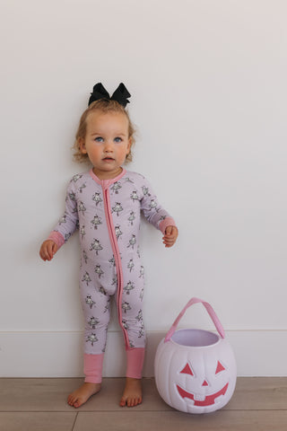 In My Jammers Ballerina Ghost Zipper Romper Child in a pink floral onesie with a black bow, standing next to a white pumpkin bucket on a light wooden floor.