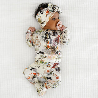 Lou Lou and Company Top and Bottoms - Nellie
A baby lying on a white textured blanket, wearing a matching long-sleeve footed sleeper and knotted headband set with a multicolor floral print.