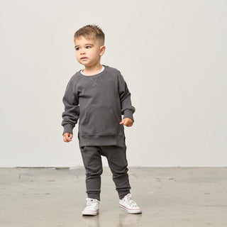 Little Bipsy Pocket Pullover - Shadow
Child wearing a gray outfit standing on a light wooden floor with a white wall background