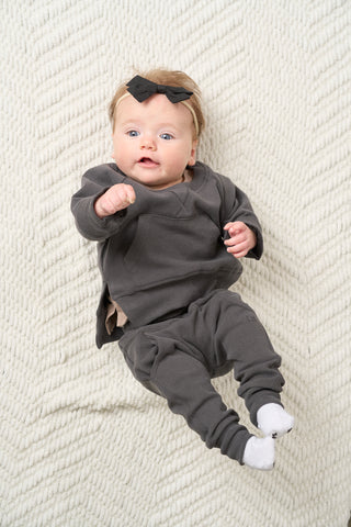 Little Bipsy Pocket Pullover - Shadow
Baby in a gray outfit with a black bow on a textured white blanket