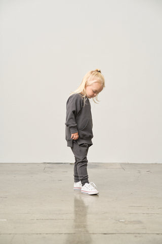 Little Bipsy Jogger - Shadow
Child wearing a gray outfit standing on a light wooden floor with a plain white wall background