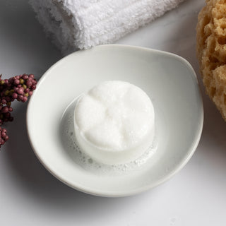 The Lavish Goat Shower Steamers Box - Essence: Eucalyptus
White bowl with a bar of soap on a light gray surface with a sponge and towel in the background.