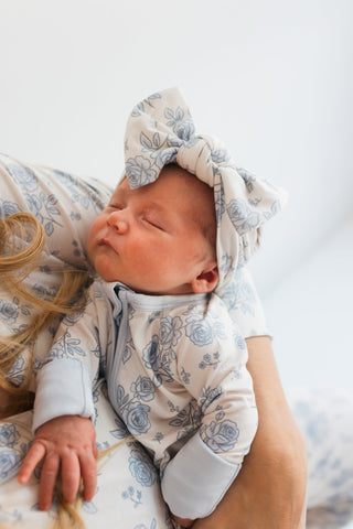 In My Jammers Blue Rose Garden Headband
Newborn baby in floral outfit and headband being held by a person against a white background