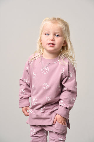 Little Bipsy Smiley Pullover - Dusty Purple
Young child wearing a pink outfit with white smiley face patterns on a plain background