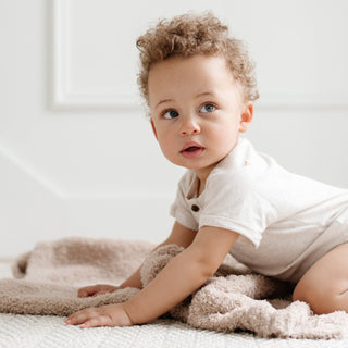Saranoni Bamboni® Receiving Blanket - Birch
Curly-haired baby in a white onesie sitting on a soft surface.