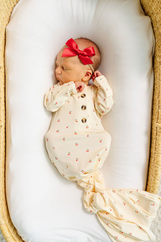Lou Lou and Company Knotted Gown - Sadie
A sleeping newborn baby wrapped in a cream-colored knit blanket with tiny red floral prints and brown buttons, wearing a matching red bow headband, lying inside a woven basket bassinet on a white cushion.