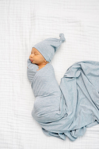 Lou Lou and Company Swaddle Blanket - Crew 
A sleeping newborn baby swaddled in a light blue muslin wrap and wearing a matching pointed hat, lying on a white textured muslin blanket.