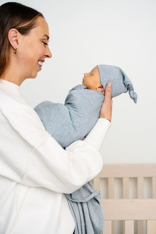 Lou Lou and Company Top Knot Hat - Crew A woman in a white long-sleeve top holds a sleeping newborn baby wrapped in a light blue heathered swaddle blanket with a matching pointed hat. The scene is set in a bright, minimalist room with a light wood crib visible in the background.