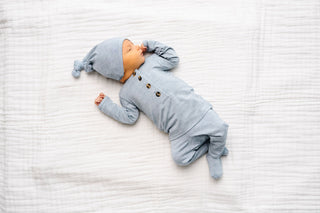 Lou Lou and Company Top and Bottoms - Crew
A sleeping newborn baby wearing a light blue footed one-piece outfit with three front buttons and a matching beanie hat, lying on a white textured muslin blanket.
