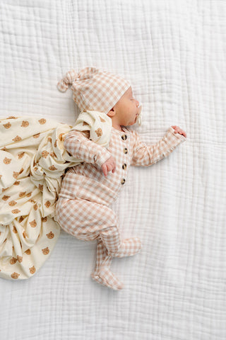 Lou Lou and Company Top and Bottoms - Milo
A newborn baby lying on a white muslin blanket, wearing a beige and white gingham footed romper and matching hat, with a cream bear-print blanket beside them.