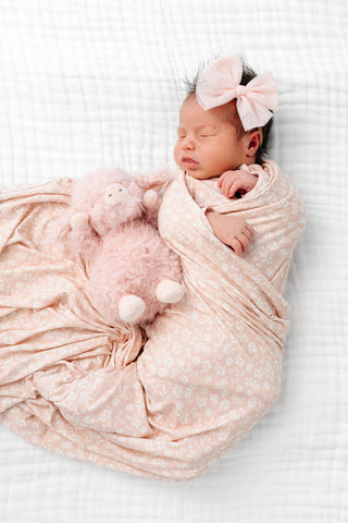 Lou Lou and Company Swaddle Blanket - Louise
A sleeping newborn baby swaddled in a light pink patterned blanket and wearing a matching pink bow headband, lying next to a plush pink stuffed toy on a white textured blanket.