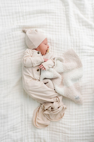 Lou Lou and Company Ribbed Knotted Gown - Cove
Newborn baby wrapped in a beige swaddle and hat, holding a plush toy on a white textured blanket.