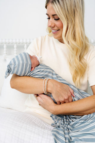 Lou Lou and Company Top Knot Hat - Wells
Woman holding a newborn baby wrapped in a striped blanket on a white bed.