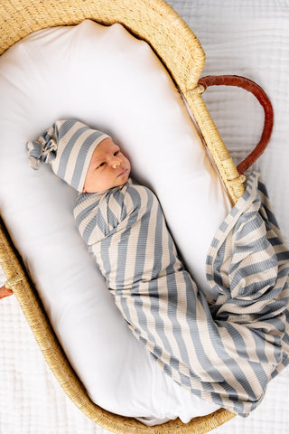 Lou Lou and Company Ribbed Swaddle Blanket - Lincoln
Newborn baby swaddled in a striped blanket and hat, lying in a wooden crib.