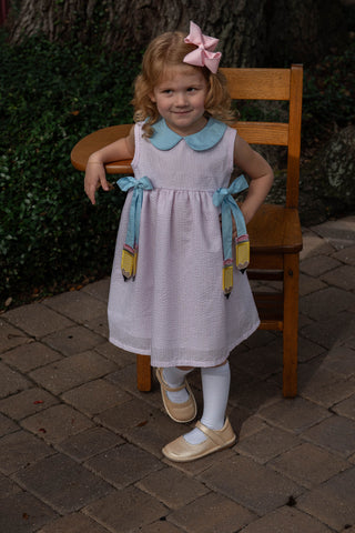 City Beautiful Pencil Dress
Young girl in a white dress with blue accents sitting on a wooden chair outdoors.