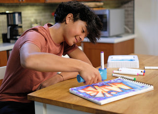 The Pencil Grip Inc Air Colors Airbrush Kit 
Person drawing in a notebook at a kitchen table with various art supplies.