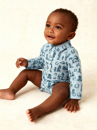 Alto Modas Winston Romper - Blue Western Toile 
A baby sitting on a cream-colored studio floor, wearing a light blue and white long-sleeve button-up shirt with a navy blue animal print.