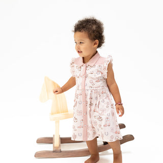 Angel Dear Western Pearl Snap Dress & Bloomer - Cowgirl Boots and Bows
A toddler girl wearing a light pink sleeveless dress with a white and pink patterned print stands next to a wooden rocking horse on a plain white background.