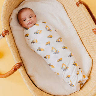 Copper Pearl Bamboo Swaddle Blanket - Dozer
A baby swaddled in a white muslin blanket with a yellow and gray construction vehicle print, lying in a woven rattan Moses basket.