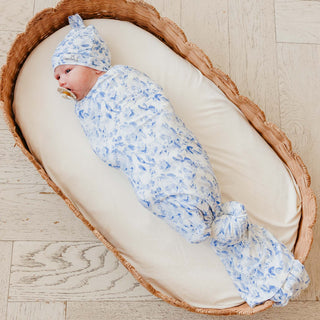 Copper Pearl Bamboo Swaddle Blanket - Priscilla
A baby swaddled in a light blue and white floral patterned swaddle blanket and matching bonnet, lying on a cream cushion inside a woven wicker bassinet on a light wood floor.