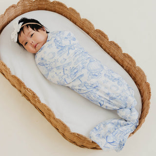 Copper Pearl Bamboo Swaddle Blanket - Vintage Carnival
A baby swaddled in a white muslin blanket with a delicate blue toile pattern, lying inside a woven rattan baby bassinet on a light cream background.