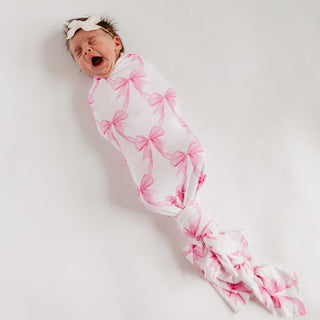 Copper Pearl Bamboo Swaddle Blanket - Bow My Gosh
A sleeping newborn baby wrapped in a white swaddle blanket with a pink bow pattern, wearing a matching white headband with a pink bow, photographed on a plain light background.